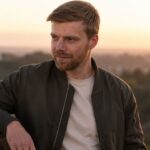 A portrait of American composer Andrew Norman looking thoughtfully into the distance against a sunset backdrop.