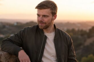 A portrait of American composer Andrew Norman looking thoughtfully into the distance against a sunset backdrop.