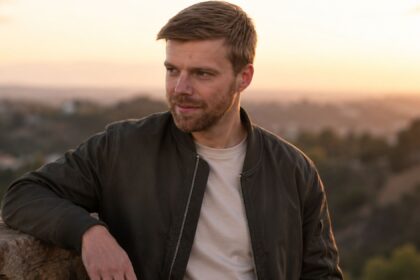 A portrait of American composer Andrew Norman looking thoughtfully into the distance against a sunset backdrop.