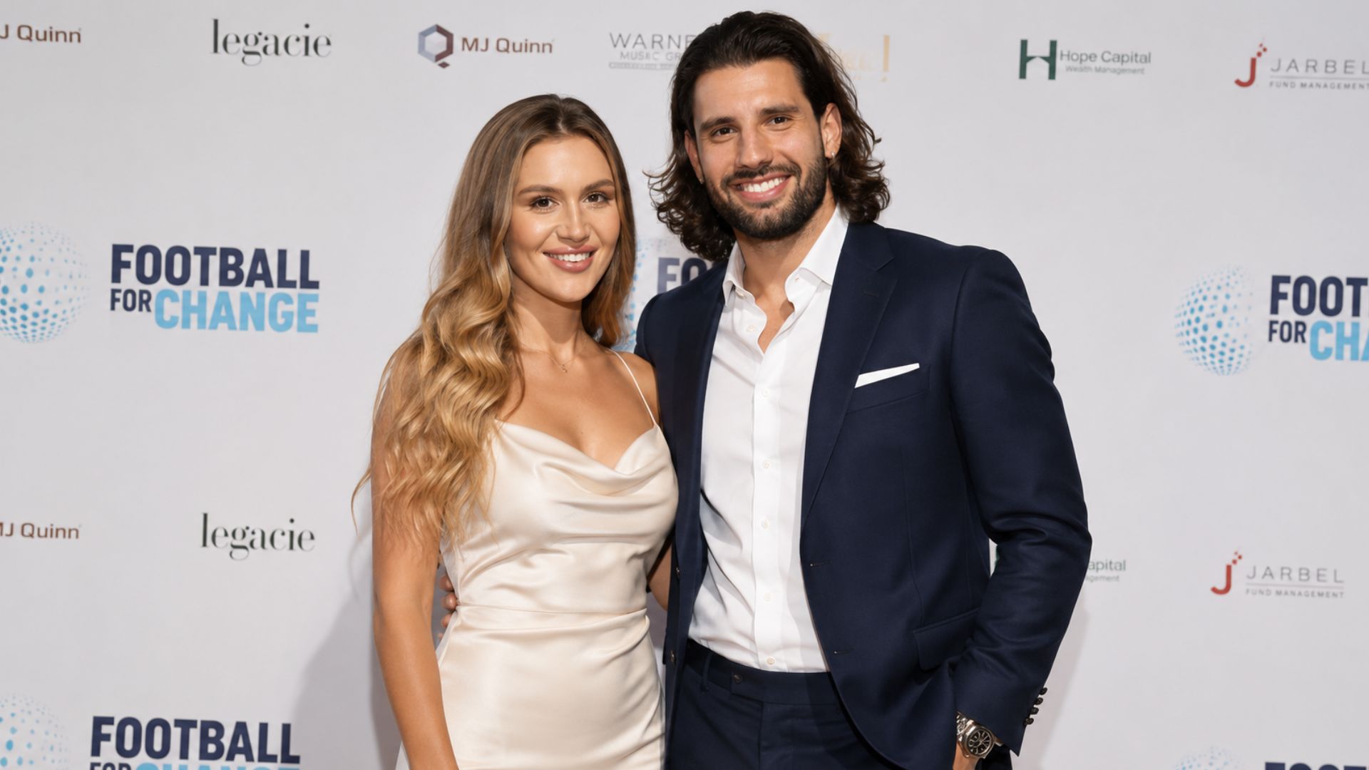 Borka Buzsik, wearing an elegant cream dress, standing alongside her husband, Liverpool footballer Dominik Szoboszlai, who is wearing a navy blue suit at a charity event.
