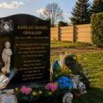 The black memorial headstone of Keelan Daniel Grealish, surrounded by bright flowers, tributes, and a small statue of a boy holding a football in a peaceful cemetery setting.