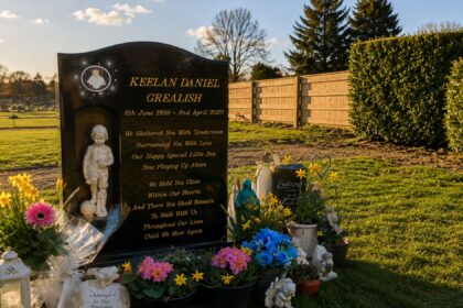 The black memorial headstone of Keelan Daniel Grealish, surrounded by bright flowers, tributes, and a small statue of a boy holding a football in a peaceful cemetery setting.