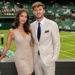 British model Kennedy Alexa, wearing a beige dress, and footballer James Maddison, in a light suit, standing together and smiling at a tennis stadium.