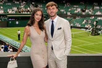 British model Kennedy Alexa, wearing a beige dress, and footballer James Maddison, in a light suit, standing together and smiling at a tennis stadium.