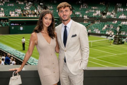 British model Kennedy Alexa, wearing a beige dress, and footballer James Maddison, in a light suit, standing together and smiling at a tennis stadium.