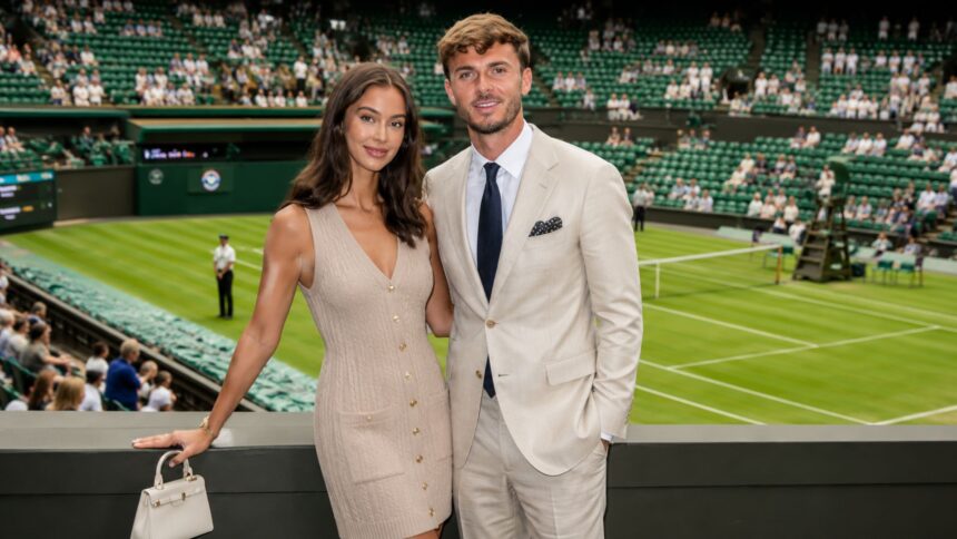British model Kennedy Alexa, wearing a beige dress, and footballer James Maddison, in a light suit, standing together and smiling at a tennis stadium.