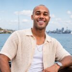 A portrait of Jules Laviscount wearing a cream-colored knit shirt, sitting outdoors with a blurred city skyline and water in the background.