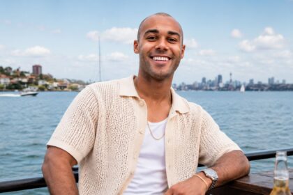 A portrait of Jules Laviscount wearing a cream-colored knit shirt, sitting outdoors with a blurred city skyline and water in the background.