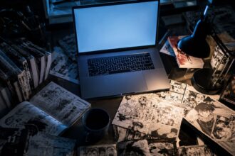 weebcentral, A dimly lit desk featuring an open laptop with a blank blue screen, surrounded by stacks of manga volumes and scattered manga pages.