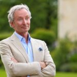 A portrait of Nigel Mackintosh, father of Millie Mackintosh, wearing a beige blazer and light blue shirt in a garden setting.
