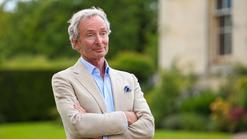 A portrait of Nigel Mackintosh, father of Millie Mackintosh, wearing a beige blazer and light blue shirt in a garden setting.