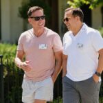 Peter Thiel, wearing a pink t-shirt, and his husband Matt Danzeisen, in a white polo shirt, smiling and talking while walking together outdoors at a conference.