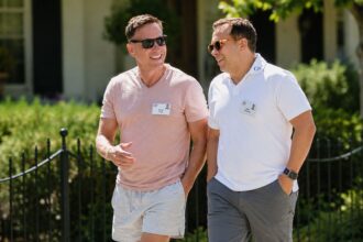 Peter Thiel, wearing a pink t-shirt, and his husband Matt Danzeisen, in a white polo shirt, smiling and talking while walking together outdoors at a conference.