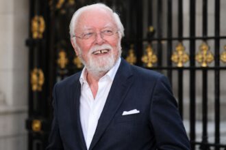 White-haired elderly man in a navy suit smiling outside ornate black gates