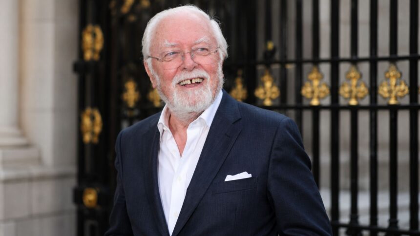 White-haired elderly man in a navy suit smiling outside ornate black gates