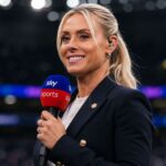 British sports broadcaster Emma Paton holding a Sky Sports microphone, smiling in a dark stadium setting.