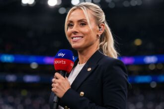 British sports broadcaster Emma Paton holding a Sky Sports microphone, smiling in a dark stadium setting.