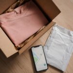 Open cardboard shipping box with folded pink sweater, prepaid return shipping label, and smartphone showing delivery tracking map