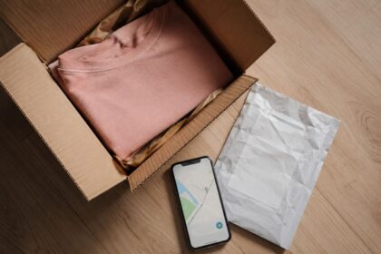 Open cardboard shipping box with folded pink sweater, prepaid return shipping label, and smartphone showing delivery tracking map