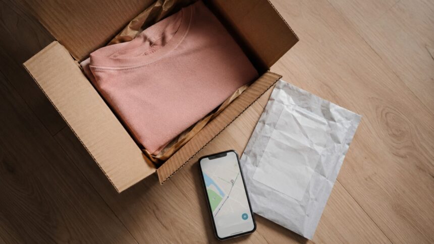 Open cardboard shipping box with folded pink sweater, prepaid return shipping label, and smartphone showing delivery tracking map