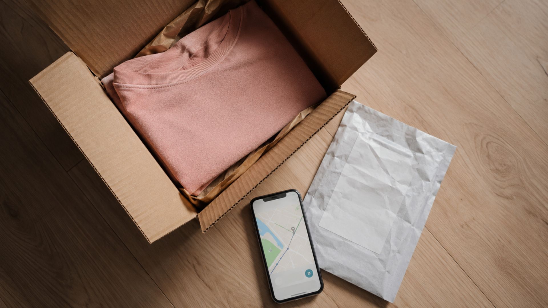Open cardboard shipping box with folded pink sweater, prepaid return shipping label, and smartphone showing delivery tracking map