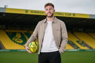 Norwich City right-back Jack Stacey standing on the pitch at Carrow Road holding a yellow and green football.