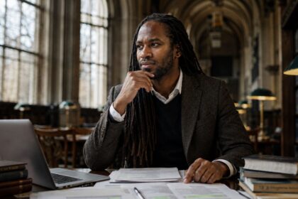 A distinguished Black British professor with long locs sits thoughtfully at a desk in a historic academic library, surrounded by books and research papers.