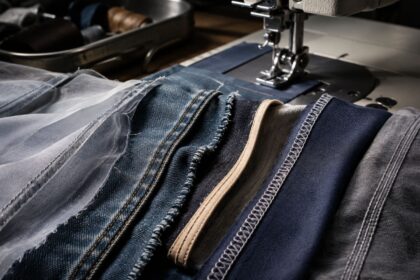 A close-up view of an industrial sewing machine needle operating over multiple fabric swatches, displaying different seam types including French, flat-felled, and overlock seams.