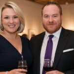 British entrepreneur Sara Davies, wearing a navy blue dress, smiling beside her husband Simon Davies, who is wearing a dark suit and purple tie, at an evening event.