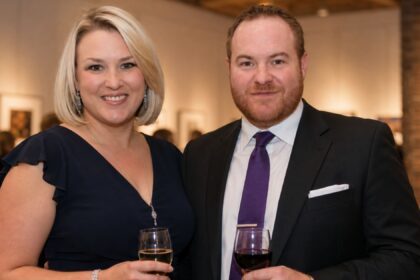British entrepreneur Sara Davies, wearing a navy blue dress, smiling beside her husband Simon Davies, who is wearing a dark suit and purple tie, at an evening event.