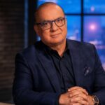 Touker Suleyman, wearing a tailored navy blue check suit and blue-rimmed glasses, sitting at a desk with a laptop against a backdrop of an evening city skyline.