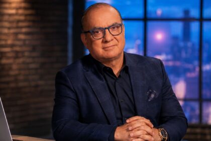 Touker Suleyman, wearing a tailored navy blue check suit and blue-rimmed glasses, sitting at a desk with a laptop against a backdrop of an evening city skyline.