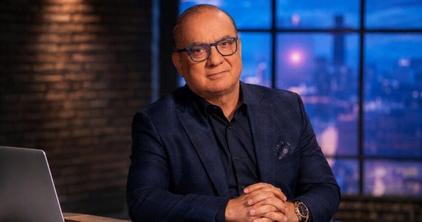 Touker Suleyman, wearing a tailored navy blue check suit and blue-rimmed glasses, sitting at a desk with a laptop against a backdrop of an evening city skyline.